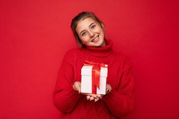 Beautiful happy young brunette woman isolated over colourful background wall wearing stylish casual clothes holding gift box and looking at camera