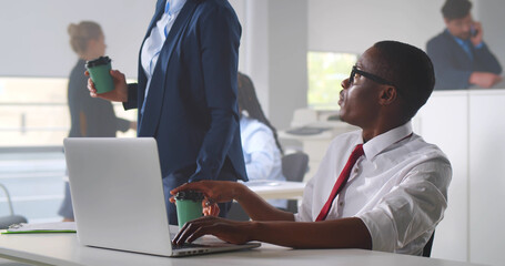 Young african businessman working with laptop at busy office and colleague bringing hom coffee