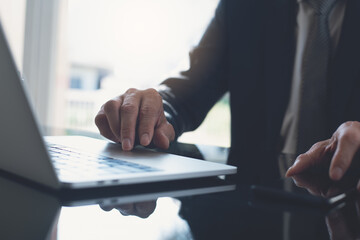 Wall Mural - Businessman in black suit working on laptop computer and using mobile smart phone at modern office