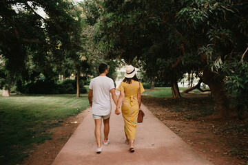 A photo from behind of a brunette girl in a yellow dress and her boyfriend who are walking on a sand path between trees in the Valencian park. A couple of tourists on a date in summer in the evening.