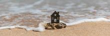 Miniature House On Natural Sea Stone Stands On Sandy Beach, Washed By Wave. Insurance, Rental, Sale Of Real Estate And Property In Seaside Resorts. Summer Holidays And Vacation Wide Banner, Copy Space