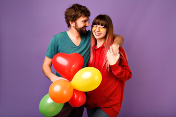 Wall Mural - Young loveble hipster couple posing on violet background, bright trendy casual clothes and glasses, hugs and smiling, friendship and relation goals, holding air balloons, ready for celebration.