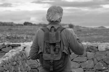 Rear view of senior man on outdoor hiking in the countryside. Backpack on his back, cloudy sky