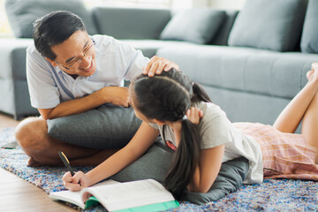 Young Asian love family father and daughter teach homework on ground floor near sofa in living room at home which smiling and felling happy. live at home concept in COVID-19 situation