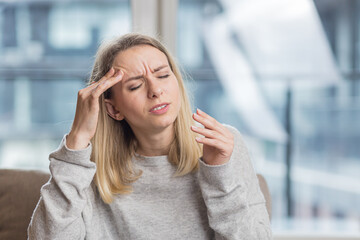 A woman holding her head anxious about an unbearable headache, sitting at home on the couch