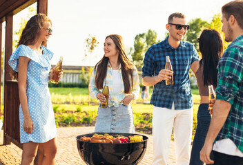 Wall Mural - Group of people standing around a barbecue, grilling outdoors, chatting and drinking drinks, outdoors in the summer in