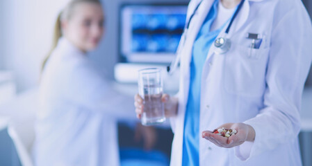 Wall Mural - shot of doctor holding pills and glass of water at clinic with colleague on the background.