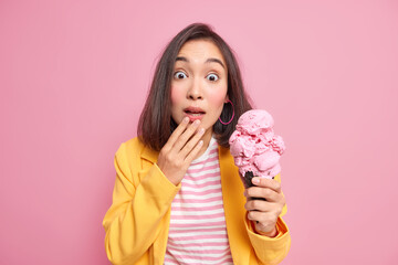 Wall Mural - Studio shot of impressed teenage girl with eastern appearance gazes surprisingly at camera holds big appetizing ice cream of raspberry flavor cannot believe her eyes dressed in elegant clothes