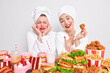 Poor nutrition concept. Happy Asian woman wears red lipstick bath towel on head eats appetizing fried chicken nuggets her friend stands near has upset expression as keeps to diet feels temptation