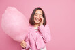 Brunette cheerful Asian woman keeps hand on face closed eyes expresses sincere feeling and emotions holds big delicious candy floss dressed formally isolated over pink background. Happiness.