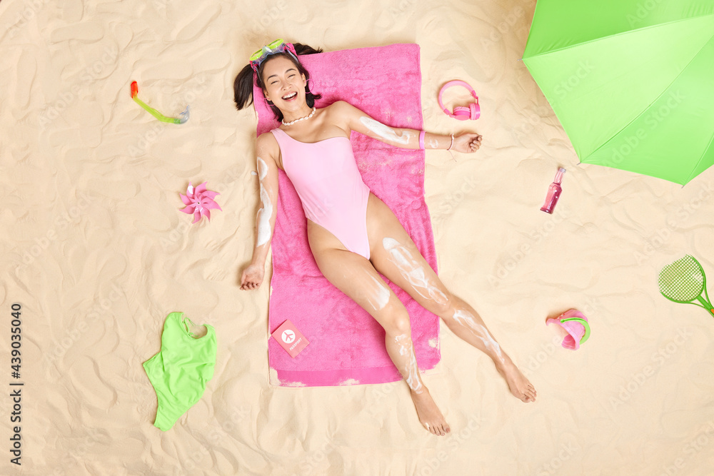 Cheerful Asian female model wears snorkeling mask pink swimsuit has upbeat mood applies suncream poses over white sand at beach surrounded by different necessary items. Summer lifestyle concept - obrazy, fototapety, plakaty 