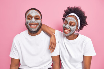 Glad Afro American couple apply clay facial mask for face care smile happily have good mood dressed in casual white t shirt spend free time together isolated over pink background. Beauty treatment