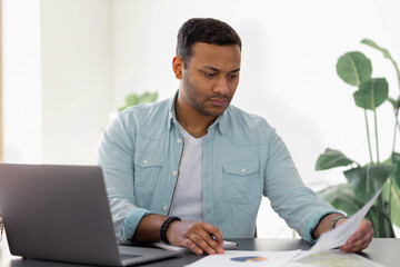 Serious young man working at home or office using laptop computer, analyzes sales reports, Indian freelancer working sitting at a desk