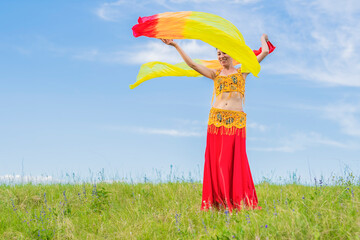 Wall Mural - A young happy woman in national costume with fiery fans in her hands is dancing an incendiary oriental dance on a green lawn against a blue sky. Outdoor activities.