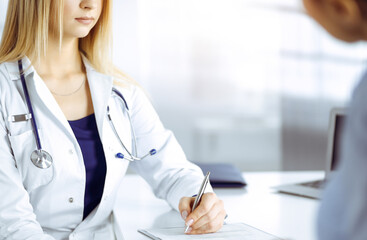 Wall Mural - Unknown woman- doctor is listening to her patient, while sitting together at the desk in the sunny cabinet in a clinic. Female physician with a stethoscope is writing at clipboard, close up. Perfect