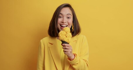 Wall Mural - Happy brunette Asian woman looks at appetizing ice cream eats tasty gelato smiles sincerely wears yellow clothes poses indoor has good mood. Eating delicious dessert. People summer food concept