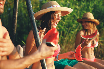 Wall Mural - Group of young people eating watermelon on a dock by the river during the summer sunny day