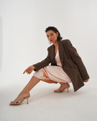 Elegant woman in peach color silk slip dress and brown squared wool blazer sitting and posing in studio at white background. Pretty stylish brunete girl with makeup and wet hair full length portrait