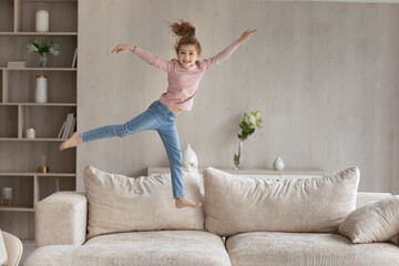 Portrait of smiling small teen biracial girl child jump on couch in living room relax alone in new renovated apartment. Happy little ethnic kid jump on sofa play at home enjoy leisure weekend.