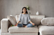 © fizkes - Calm Latino woman sit relax on sofa at home meditate practice yoga with mudra hands. Peaceful Hispanic mom rest on couch relieve stress negative emotions. Hobby, peace, meditation concept.