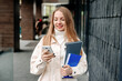 © Evgenia - portrait of blonde female student smiling and using mobile phone, texting, typing and reading message against college building background