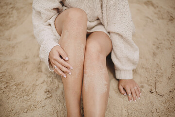 Beautiful woman with tanned legs in sand sitting on sandy beach with green grass, close up. Carefree tranquil moment. Stylish young sexy female in sweater relaxing on coast