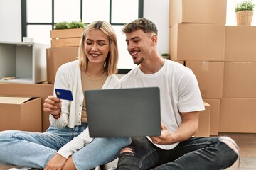 Canvas Print - Young caucasian couple smiling happy using laptop and credit card sitting at new home.