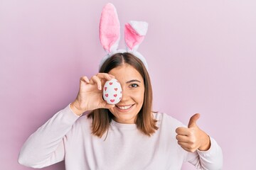 Canvas Print - Young beautiful woman wearing cute easter bunny ears holding egg smiling happy and positive, thumb up doing excellent and approval sign