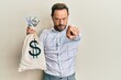 © Krakenimages.com - Middle age man wearing business shirt holding dollars bag pointing with finger to the camera and to you, confident gesture looking serious