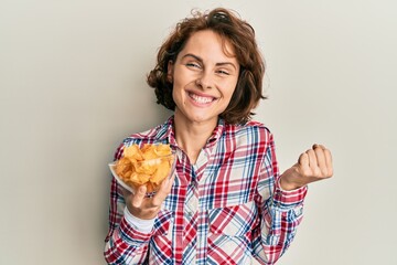 Poster - Young brunette woman holding potato chips screaming proud, celebrating victory and success very excited with raised arm