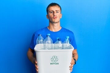 Young caucasian man holding recycling wastebasket with plastic bottles looking at the camera blowing a kiss being lovely and sexy. love expression.