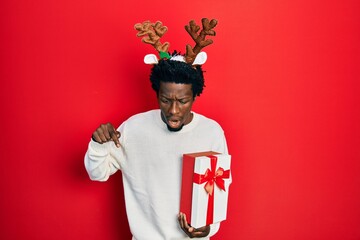 Poster - Young african american man wearing deer christmas hat holding gift pointing down with fingers showing advertisement, surprised face and open mouth