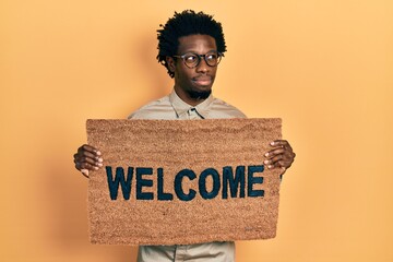 Sticker - Young african american man holding welcome doormat smiling looking to the side and staring away thinking.