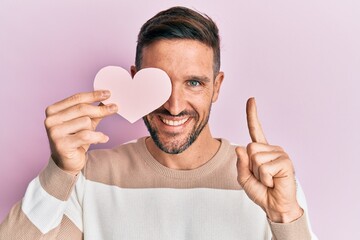 Sticker - Handsome man with beard holding heart shape paper smiling with an idea or question pointing finger with happy face, number one