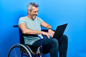 Sticker - Handsome middle age man with grey hair sitting on wheelchair using laptop winking looking at the camera with sexy expression, cheerful and happy face.