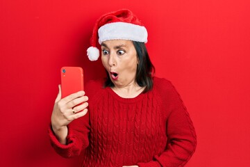 Poster - Middle age hispanic woman wearing christmas hat having video call using smartphone scared and amazed with open mouth for surprise, disbelief face