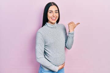 Canvas Print - Beautiful woman with blue eyes expecting a baby, touching pregnant belly pointing to the back behind with hand and thumbs up, smiling confident