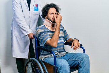 Sticker - Handsome hispanic man sitting on wheelchair wearing neck collar smelling something stinky and disgusting, intolerable smell, holding breath with fingers on nose. bad smell