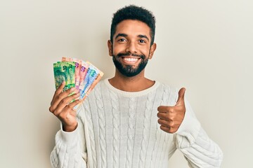 Poster - Handsome hispanic man with beard holding south african rand banknotes smiling happy and positive, thumb up doing excellent and approval sign