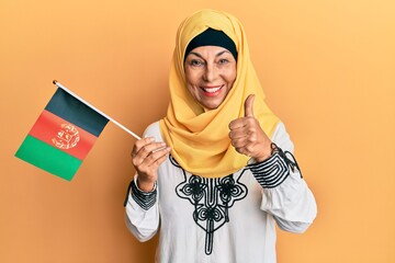 Sticker - Middle age hispanic woman holding afghanistan flag smiling happy and positive, thumb up doing excellent and approval sign