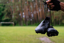Black Football Boots In Men's Hands On A Football Field Background. Raindrops On Shoes. Forest Stadium After The Rain In Ukraine. Copy Space.