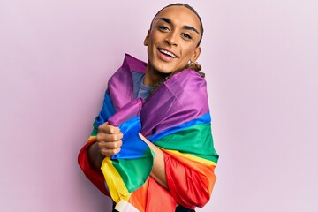 Hispanic man wearing make up and long hair wrapped in rainbow lgbtq flag celebrating crazy and amazed for success with open eyes screaming excited.