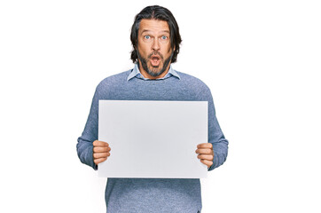 Middle age handsome man holding blank empty banner scared and amazed with open mouth for surprise, disbelief face