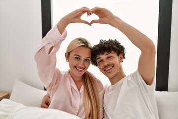 Wall Mural - Young beautiful couple doing heart symbol with hands sitting on the bed at home.