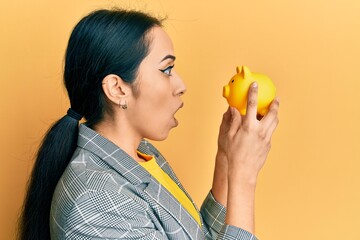 Wall Mural - Young hispanic girl looking to piggy bank afraid and shocked with surprise and amazed expression, fear and excited face.