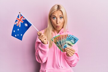 Poster - Young blonde woman holding australian flag and dollars in shock face, looking skeptical and sarcastic, surprised with open mouth