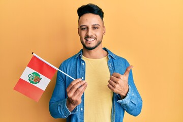 Young arab man holding peru flag smiling happy and positive, thumb up doing excellent and approval sign