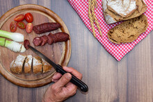 A Bavarian Snack On An Old Rustic Wooden Board With Sausage, Tomatoes And Onions, Next To It A Red White Cloth With Bread On A Table. A Hand With A Knife Takes A Piece Of Sausage