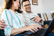 © Zoran Zeremski - Smiling couple sitting on sofa at home using laptop for shopping online.