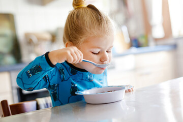 Gorgeous little toddler girl eating healthy cereal with milk for breakfast. Cute happy baby child in colorful clothes sitting in kitchen and having fun with preparing oats, cereals. Indoors at home